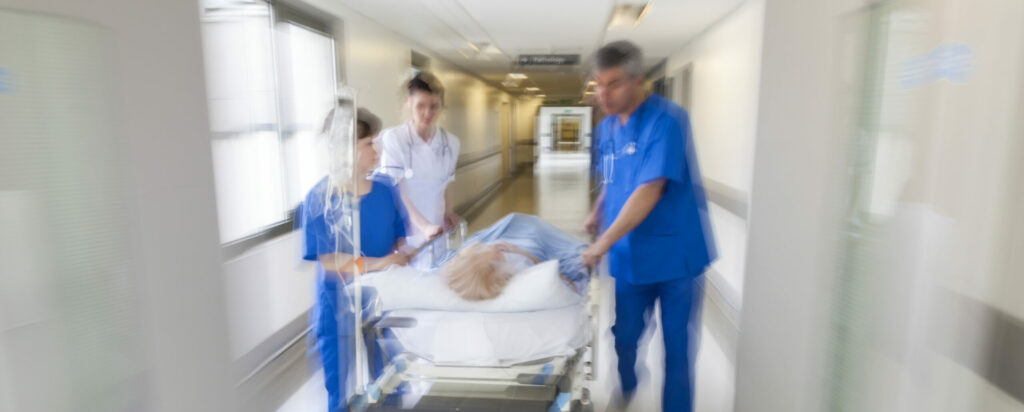 A motion blurred photograph of a patient on stretcher or gurney being pushed at speed through a hospital corridor by doctors and nurses to an emergency room